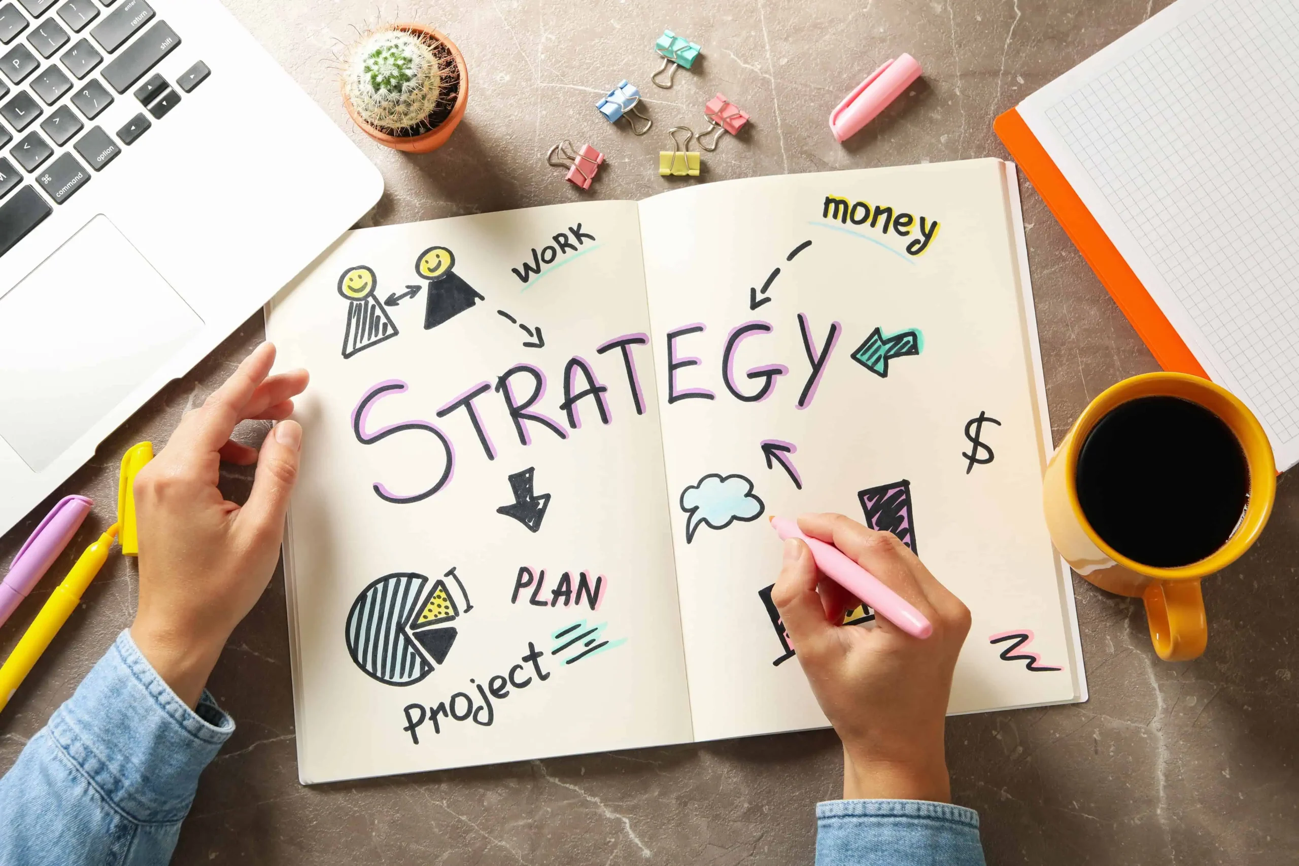 Mano escribiendo en una libreta la palabra “strategy” con dibujos relacionados a planificación, dinero y trabajo, acompañada de un café, una laptop y útiles de oficina.