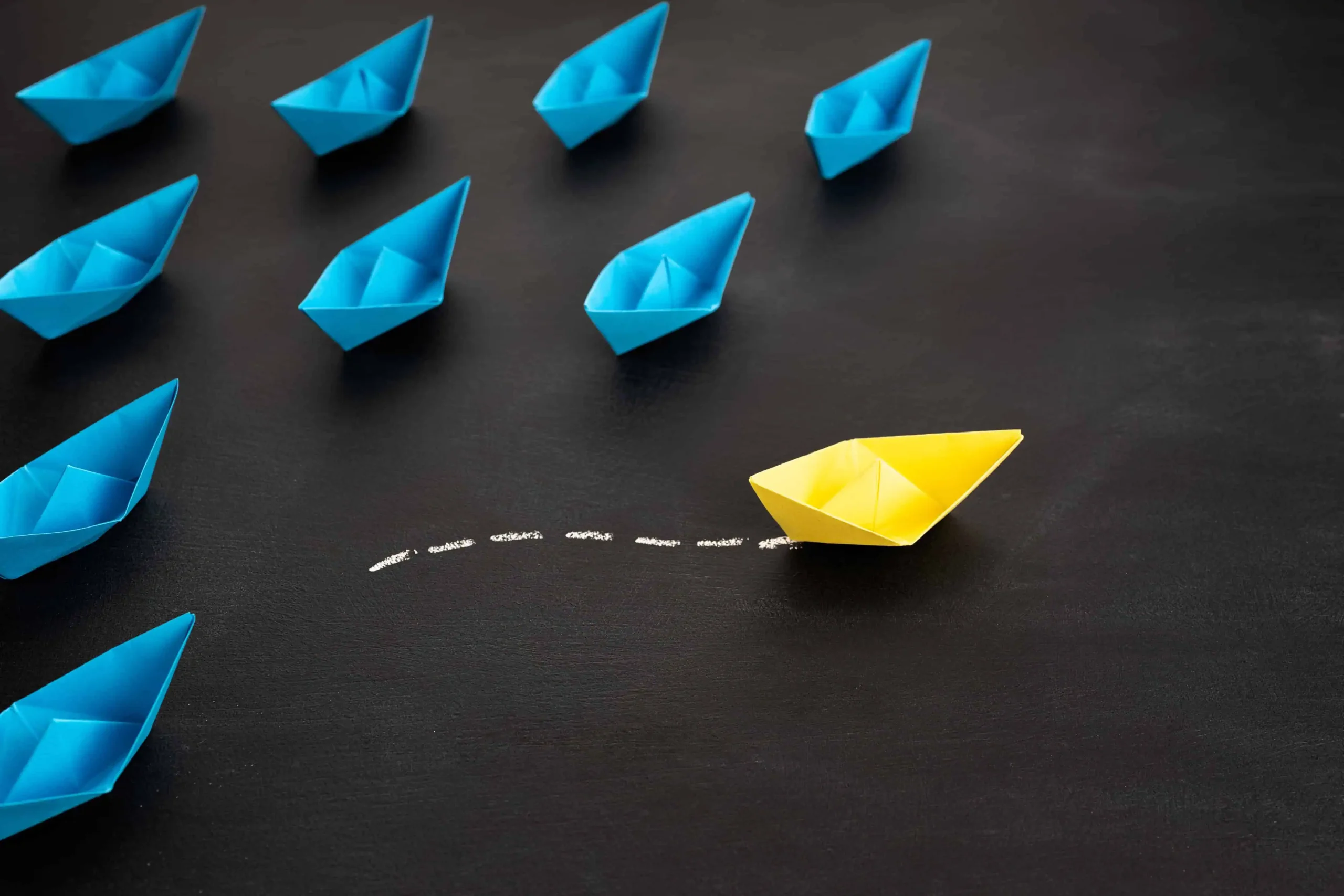 Barco de papel amarillo desviándose del grupo de barcos azules sobre fondo negro, simbolizando liderazgo para emprendedores e innovación.