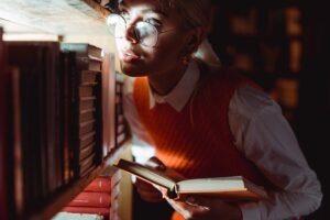 Joven con gafas y suéter naranja leyendo un libro en una biblioteca con luz tenue