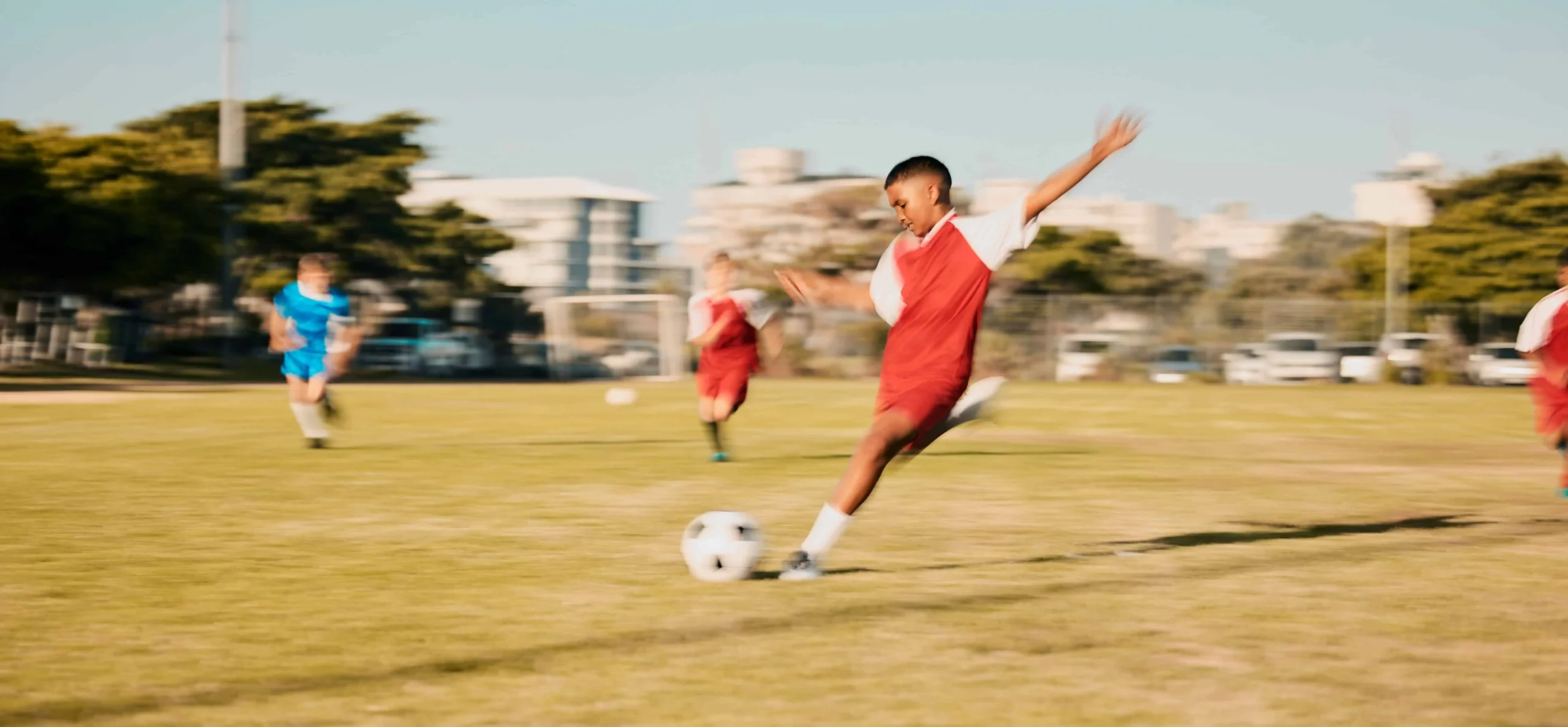 Niño con uniforme rojo y blanco pateando un balón de fútbol en un campo de césped, con jugadores en el fondo.